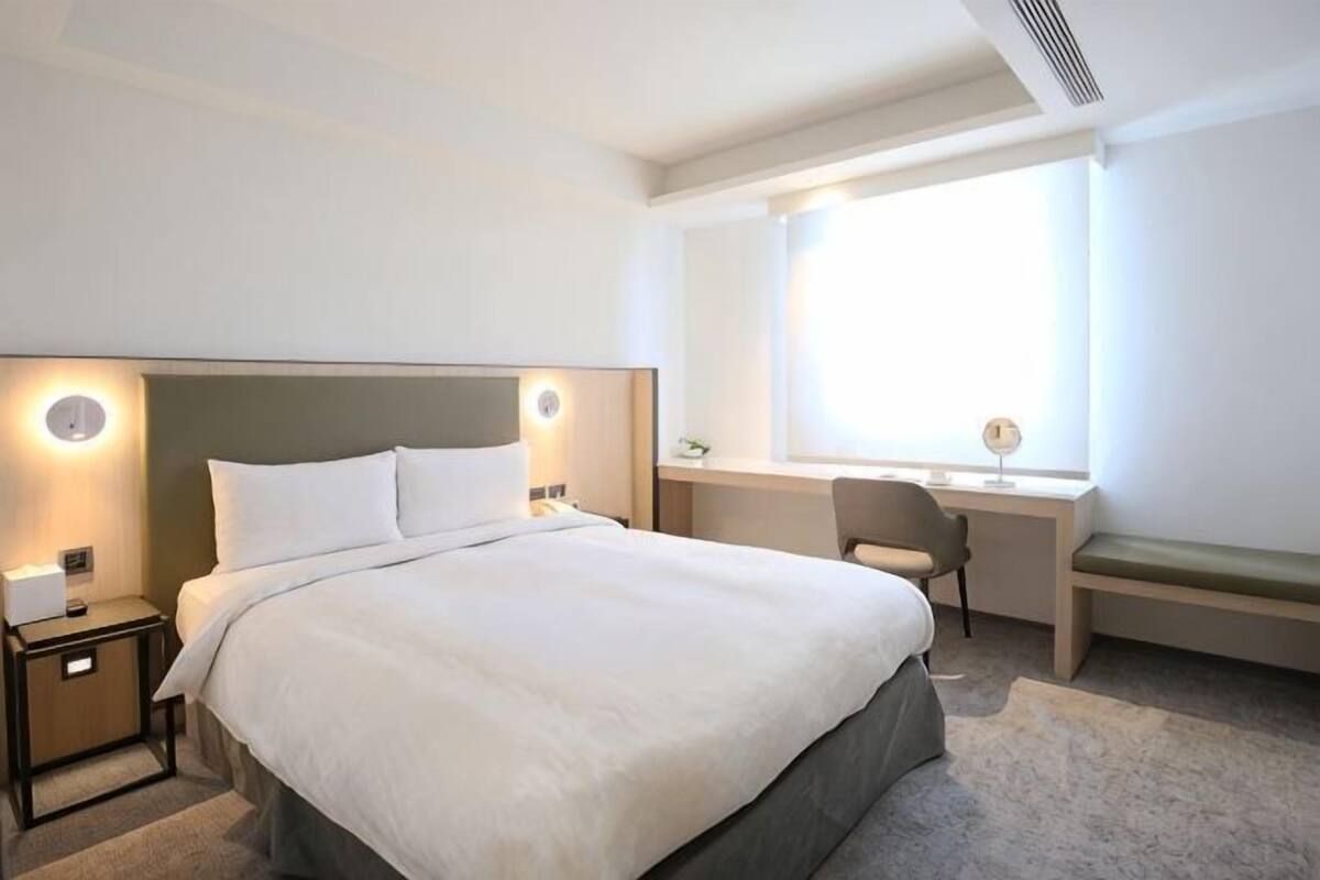 Quarto de hotel com uma cama grande, mesas de cabeceira, uma escrivaninha, uma cadeira e uma janela.
