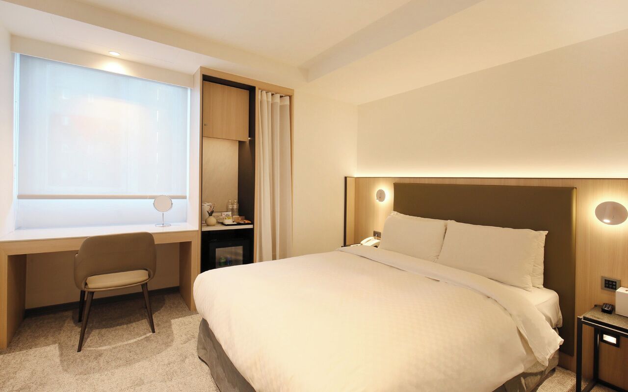 A hotel room with a bed, a desk, a chair, a nightstand, and a window with curtains.