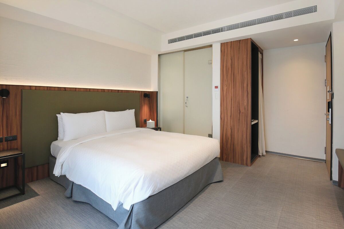 A modern hotel room with a large bed, a wooden headboard, a walk-in closet, and a bedside table.