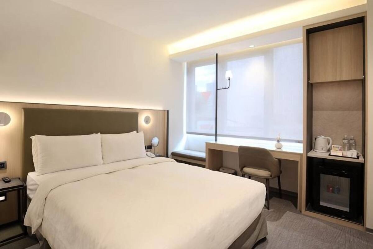 Quarto de hotel com cama, mesa de cabeceira, escrivaninha, cadeira e um pequeno espaço de cozinha.