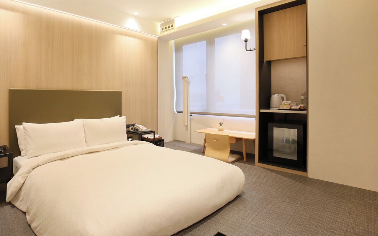 A hotel room with a large bed, a desk, a small kitchenette, and a window with blinds.