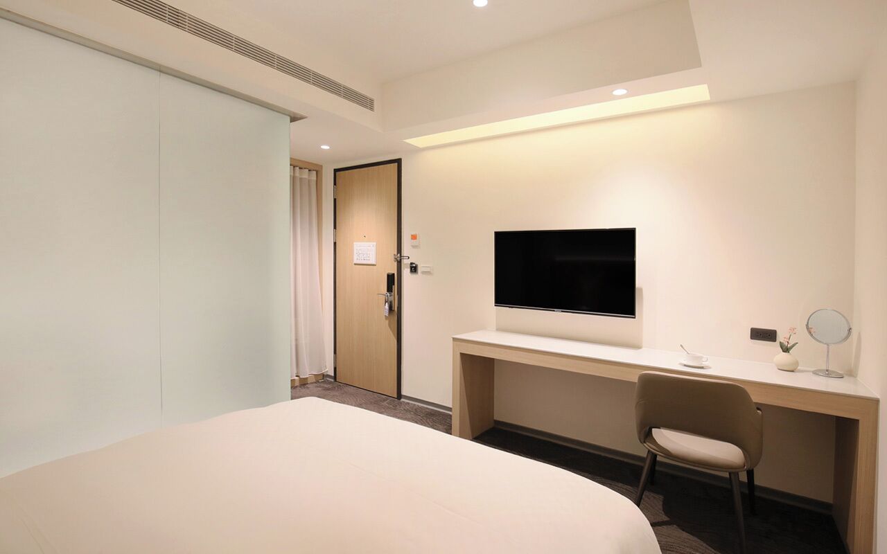 A hotel room with a bed, a desk, a chair, a TV, and a door.