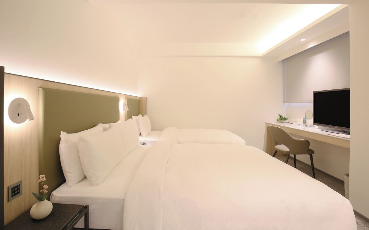 A modern hotel room with a large bed, a desk with a flat-screen TV, and a small table with a vase of flowers.