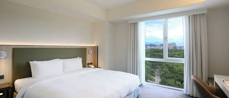 A hotel room with a large bed, a desk, and a view of a cityscape through a large window.