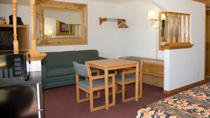 A cozy room with a wooden-framed mirror, a green sofa, a wooden table with two chairs, and a bed with a patterned bedspread.