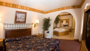 A bedroom with a bed, bedside lamps, a TV, a desk, a plant, and a bathroom visible through an archway.