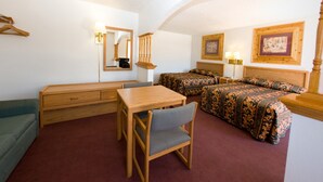 A hotel room with a wooden desk, two single beds with patterned bedding, a green sofa, a mirror, and a framed picture on the wall.