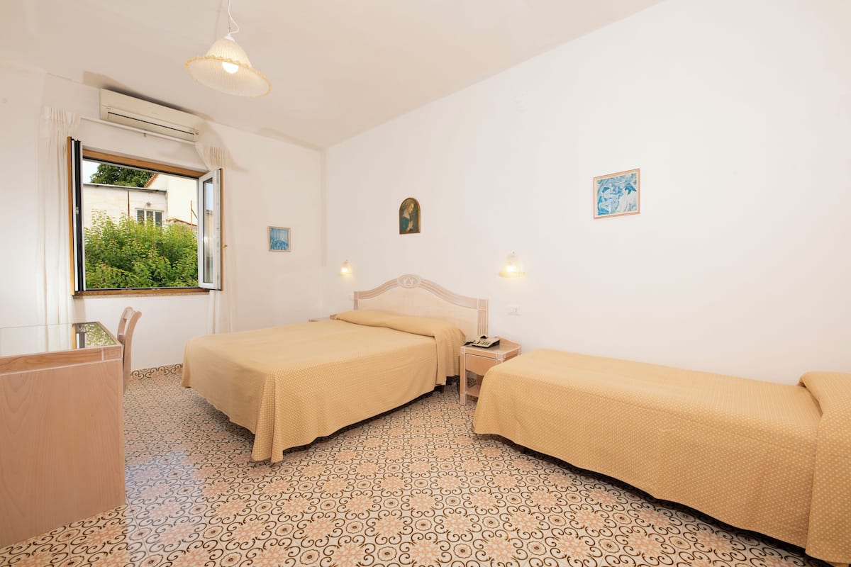 A room with two beds, a wooden desk, a window with a view of greenery, and patterned floor tiles.
