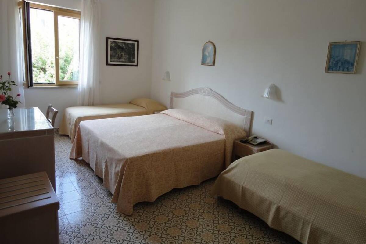 A bedroom with two single beds, a desk, and a chair. There is a window with curtains, a wall-mounted lamp, and a framed picture.