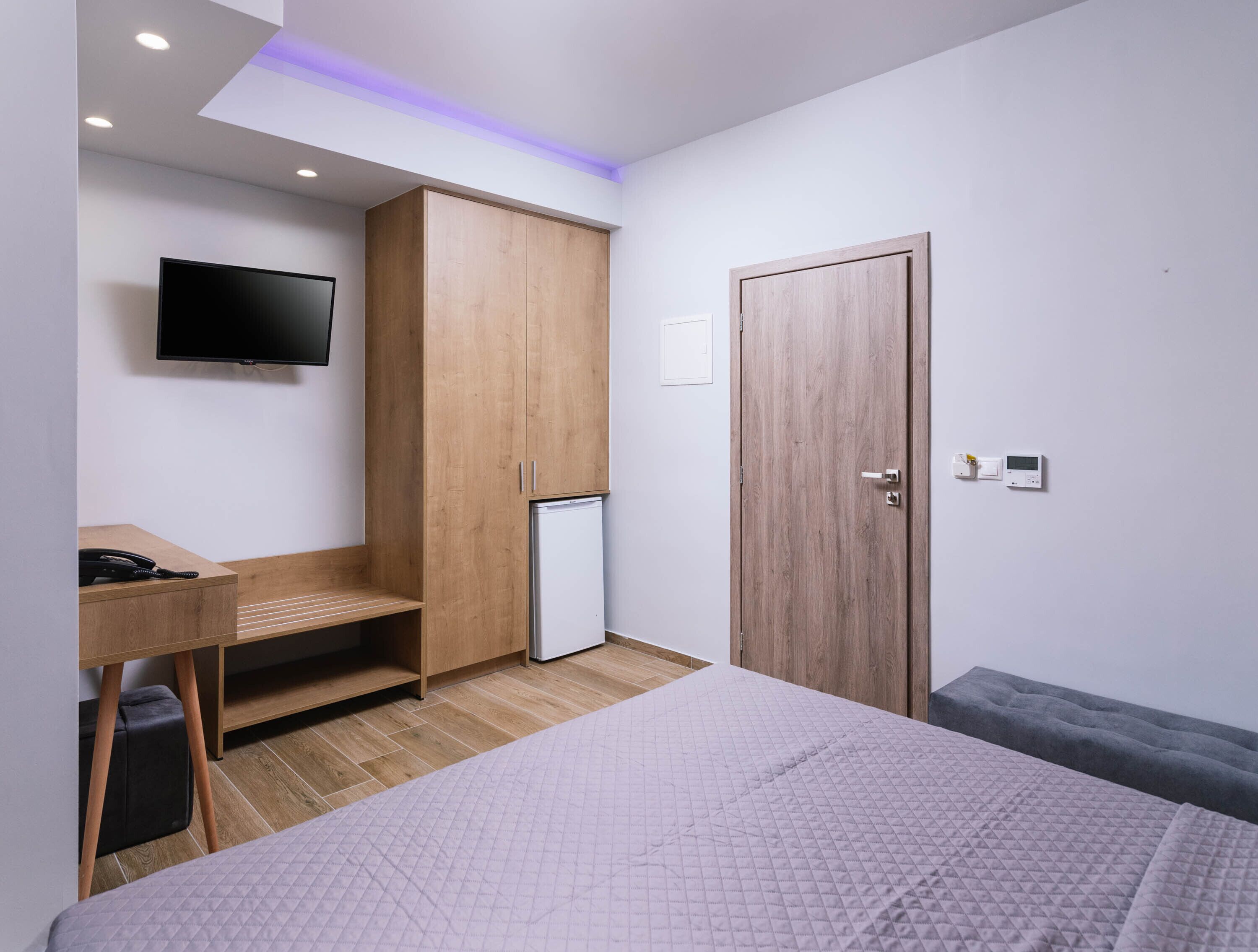 A modern bedroom with a bed, a desk, a wardrobe, a television, and a mini-fridge.