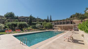 A swimming pool with lounge chairs, a stone wall, and a view of a hillside with trees and buildings.