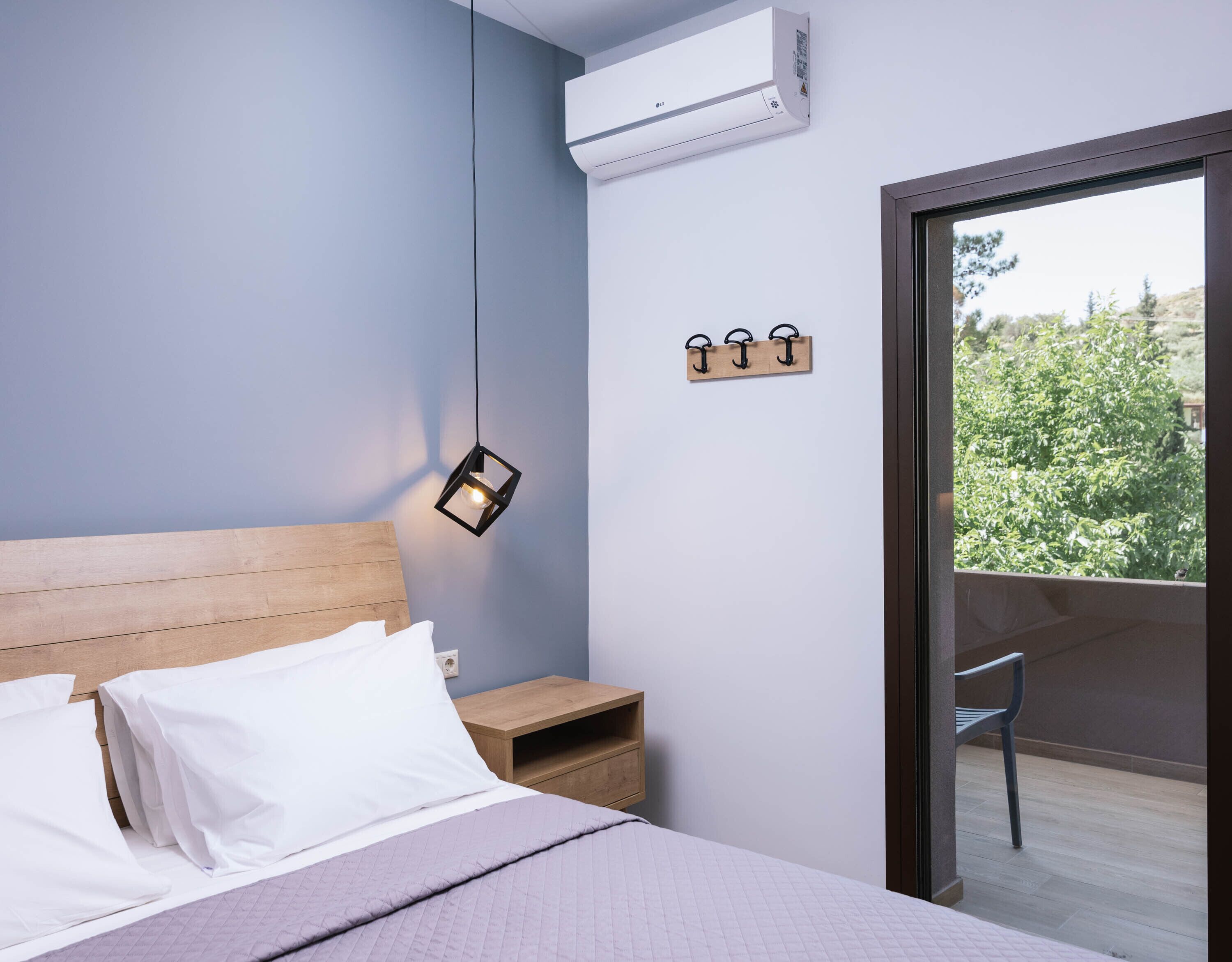 A bedroom with a bed, bedside table, nightstand, and a view of greenery through a balcony door.