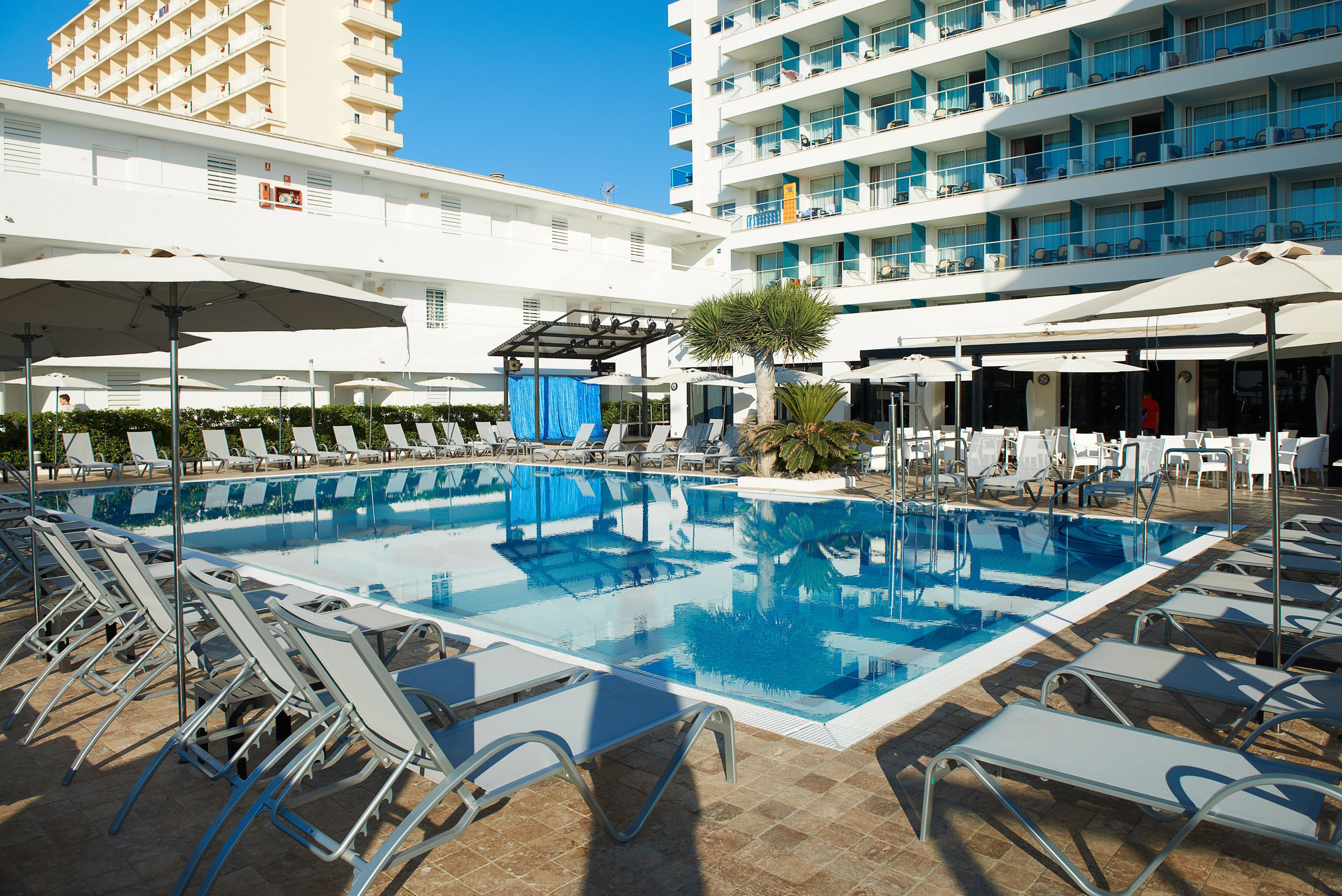 A rooftop pool area with sun loungers and umbrellas, surrounded by a multi-story hotel building.