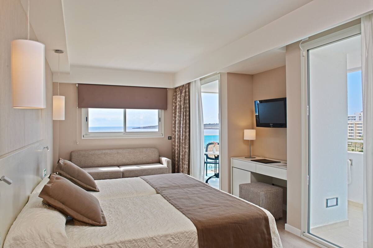 Ein Hotelzimmer mit Bett, einer Sitzecke, einem Schreibtisch mit Fernseher und einem Balkon mit Meerblick.
