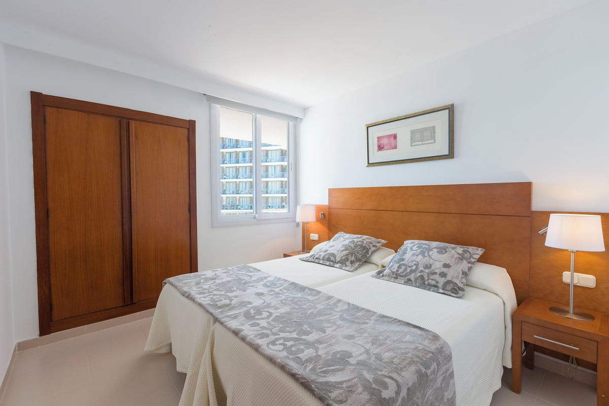 Un dormitorio con una cama grande, un armario de madera, una mesita de noche con lámpara y una ventana con vista a edificios.