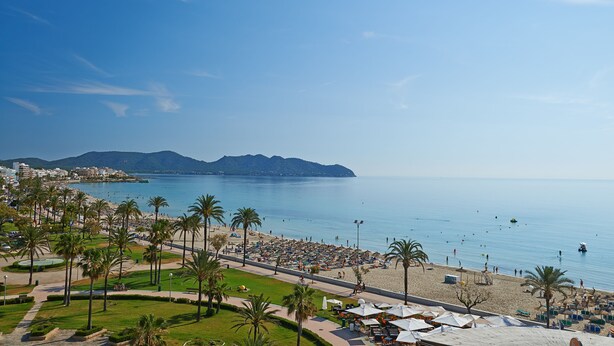 Blick auf die Küste mit Strand, Palmen und einer Stadtlandschaft im Hintergrund.