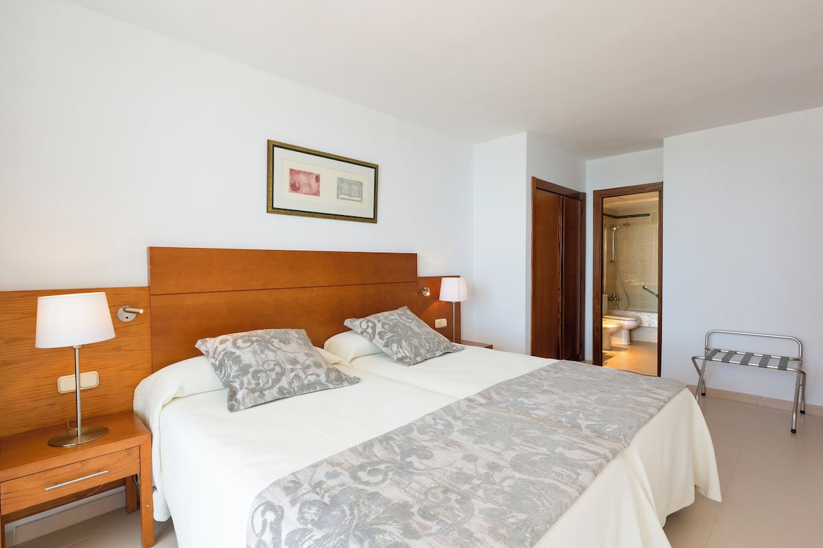 Un dormitorio ordenado con una cama, mesitas de noche, un cuadro enmarcado y un baño visible a través de una puerta abierta.