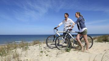 Escena de playa con dos personas en bicicletas, una sosteniendo a la otra, cerca del océano.