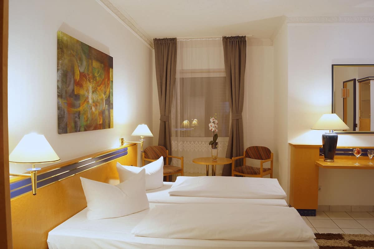 A hotel room with two beds, a desk, a chair, a small table, a lamp, a painting, and a window with curtains.