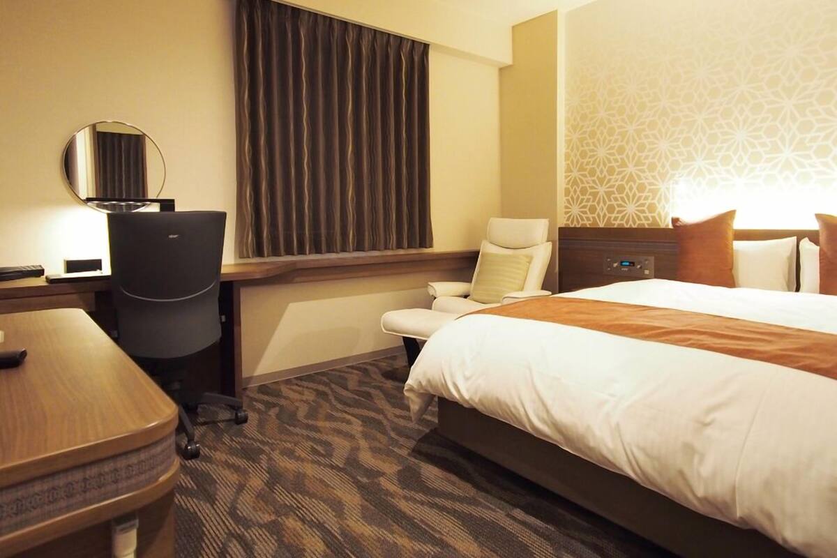 Ein Hotelzimmer mit Bett, Schreibtisch, Stuhl und einem großen Fenster mit Vorhängen.