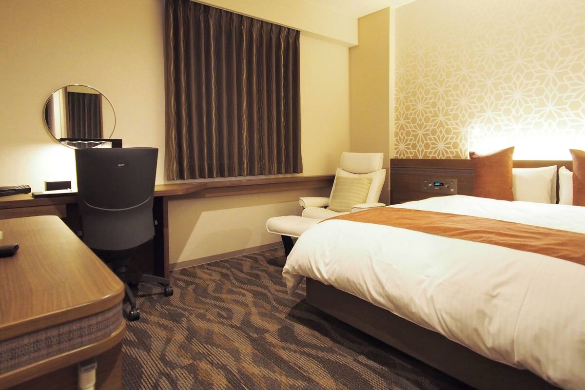 A hotel room with a bed, a desk with a chair, a small table, a mirror, and a window with curtains.