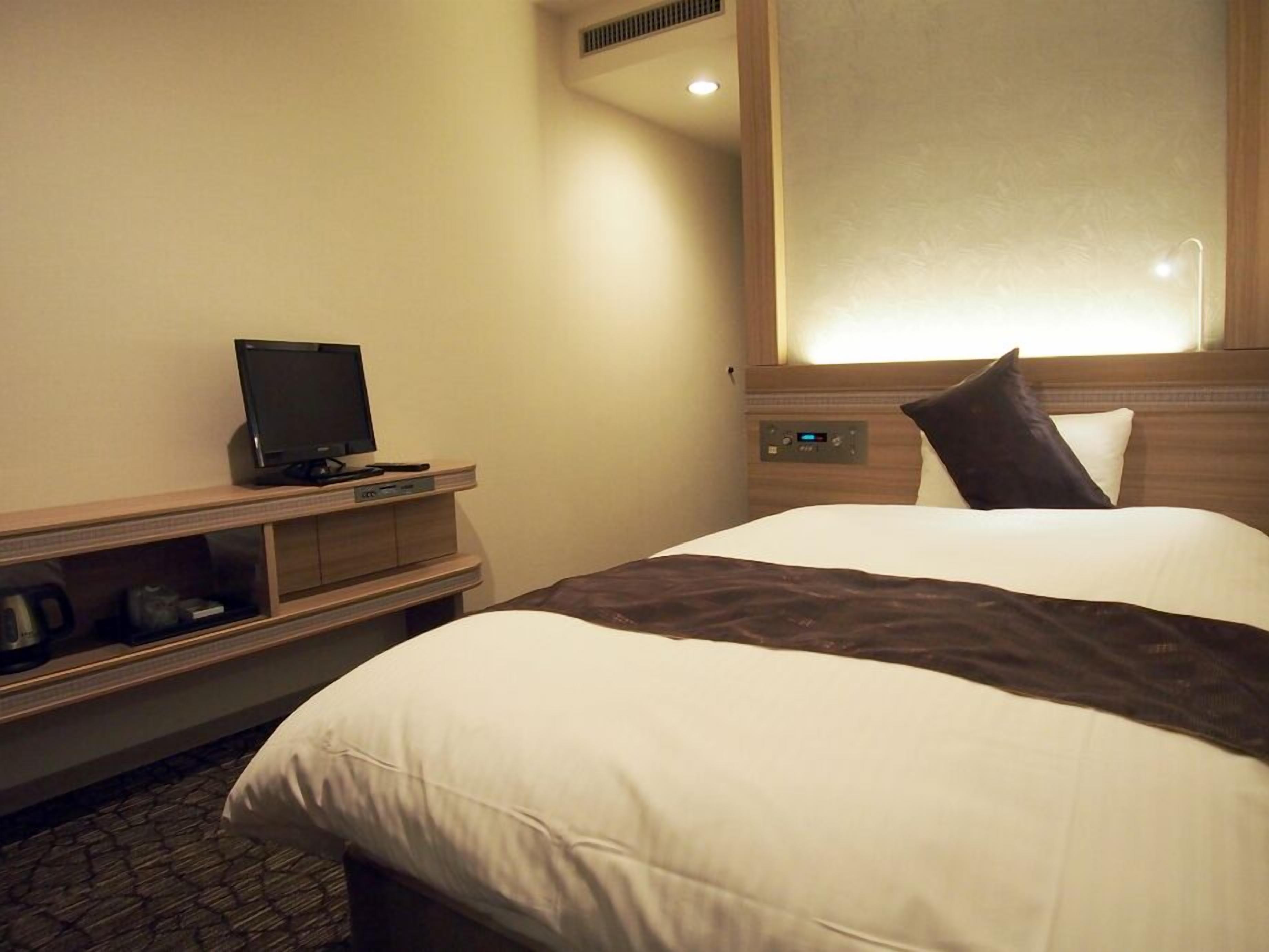 Ein Hotelzimmer mit Bett, Fernseher auf einem Holztisch, Bedienfeld und Fenster mit Rollläden.