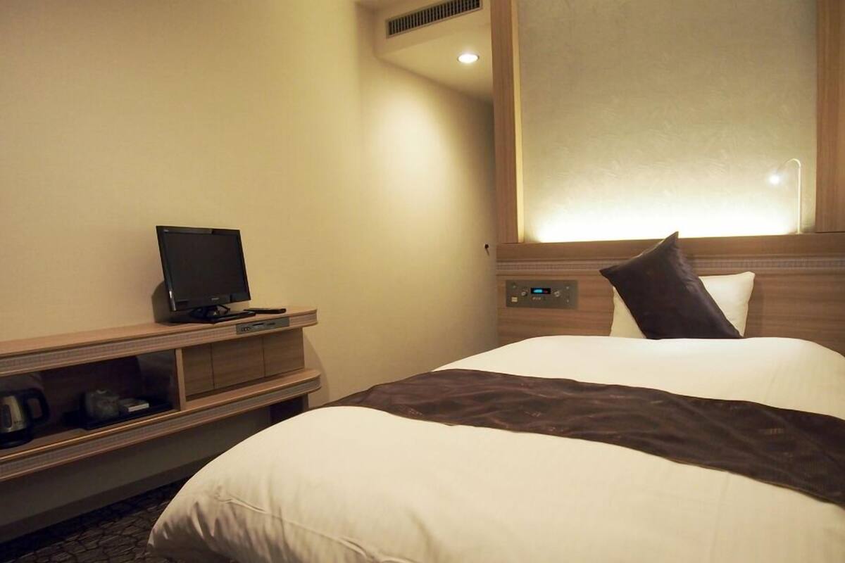 A hotel room with a bed, a television on a wooden stand, a control panel, and a window with blinds.