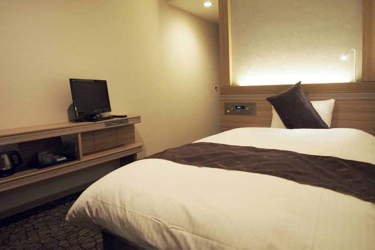 A hotel room with a bed, a television on a wooden stand, and a control panel on the wall.