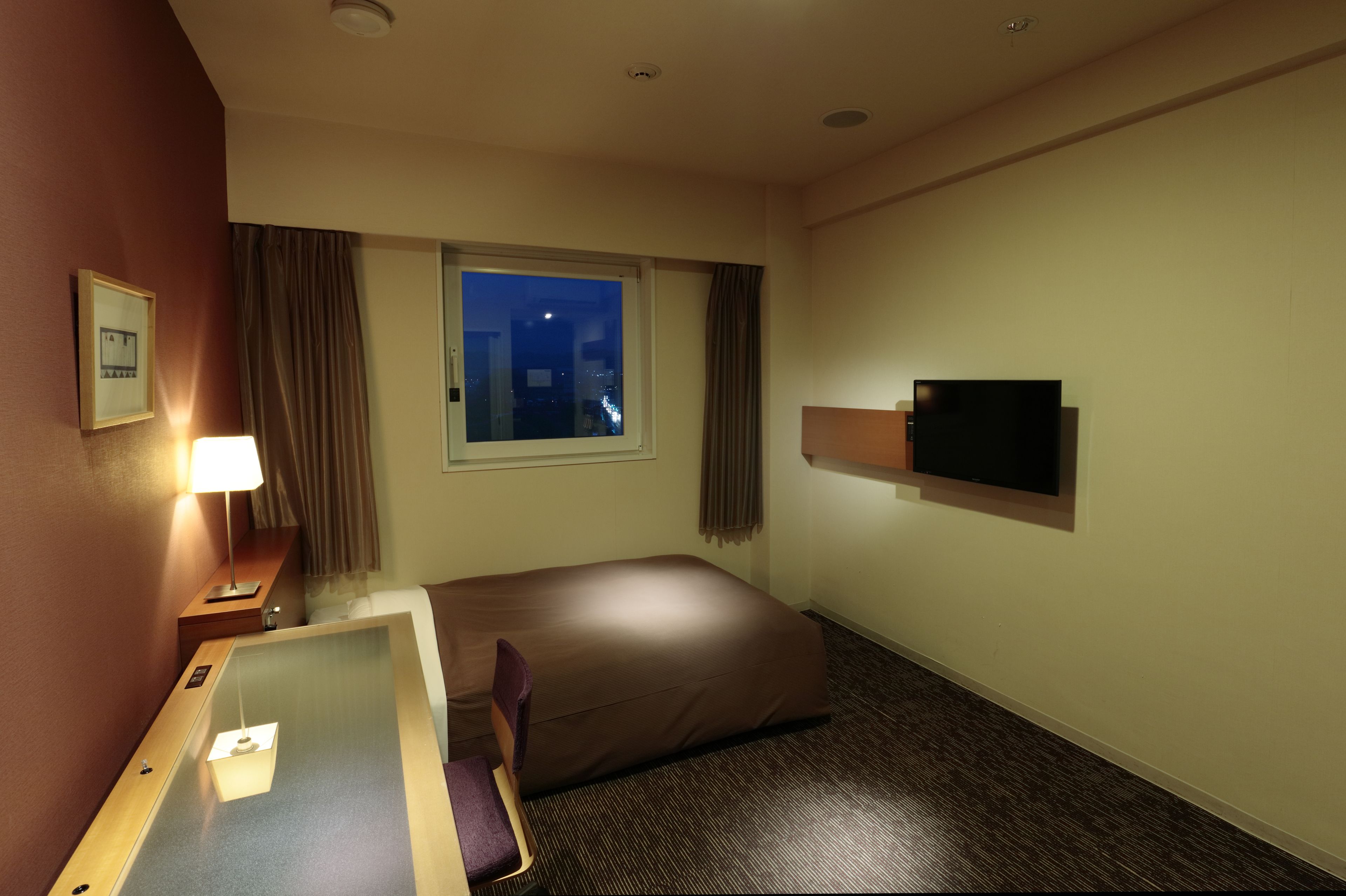 A hotel room with a bed, a desk, a television, a window with a city view, and a night sky visible through the window.