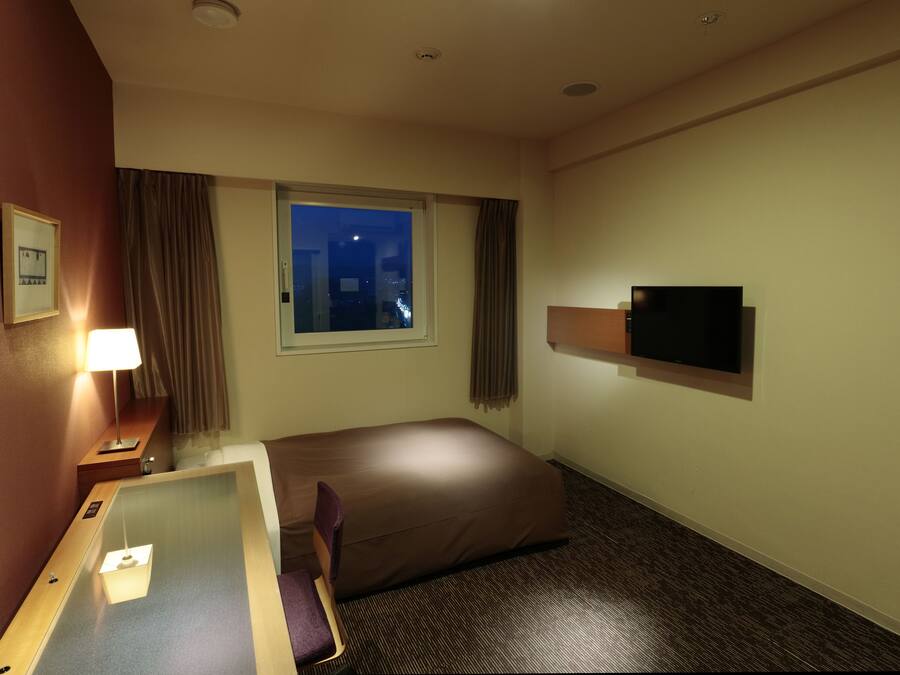 A hotel room with a bed, a desk, a television, a window with a city view, and a night sky visible through the window.