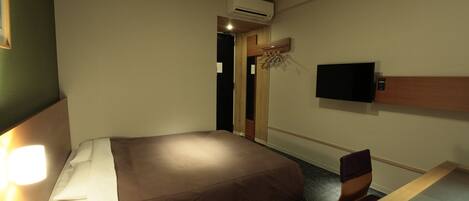 A hotel room with a bed, a desk, a chair, a TV mounted on the wall, and an air conditioning unit.