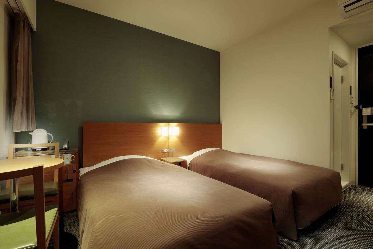 A hotel room with two single beds, a wooden headboard, a small table with a kettle, and a chair.