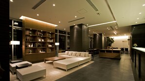 A modern hotel lobby with a sofa, ottoman, and wooden shelving with bottles and framed pictures. - Candeo Hotels Kameyama (Kameyama)