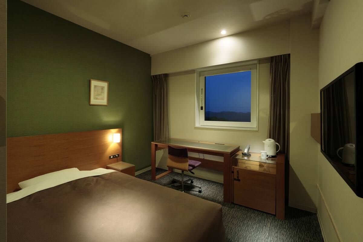 A hotel room with a bed, desk, chair, and a window with a view of the mountains.