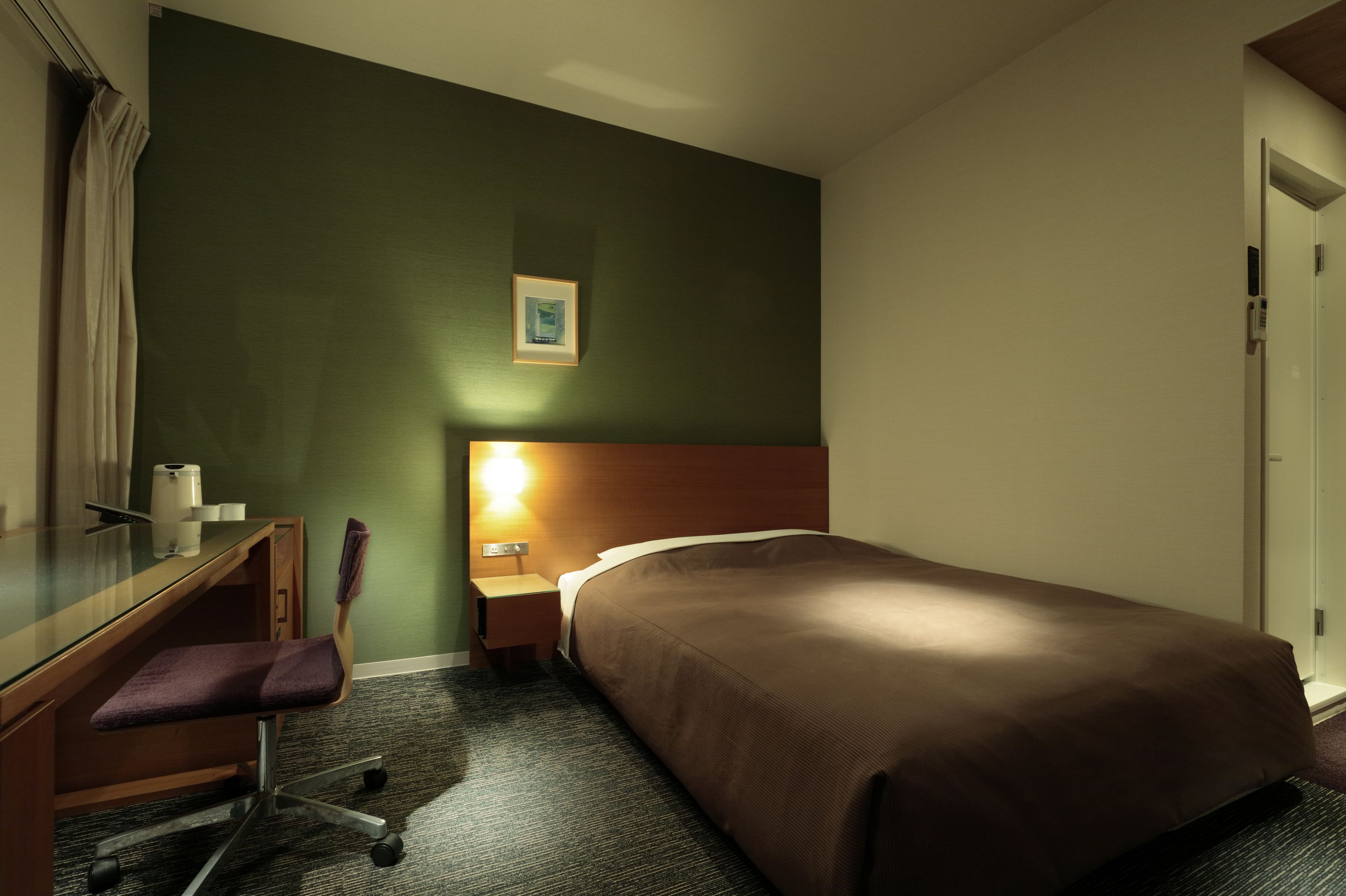 Ein Hotelzimmer mit einem Bett, einem Schreibtisch mit Stuhl, einem Nachttisch mit Lampe und einem gerahmten Bild an der Wand.