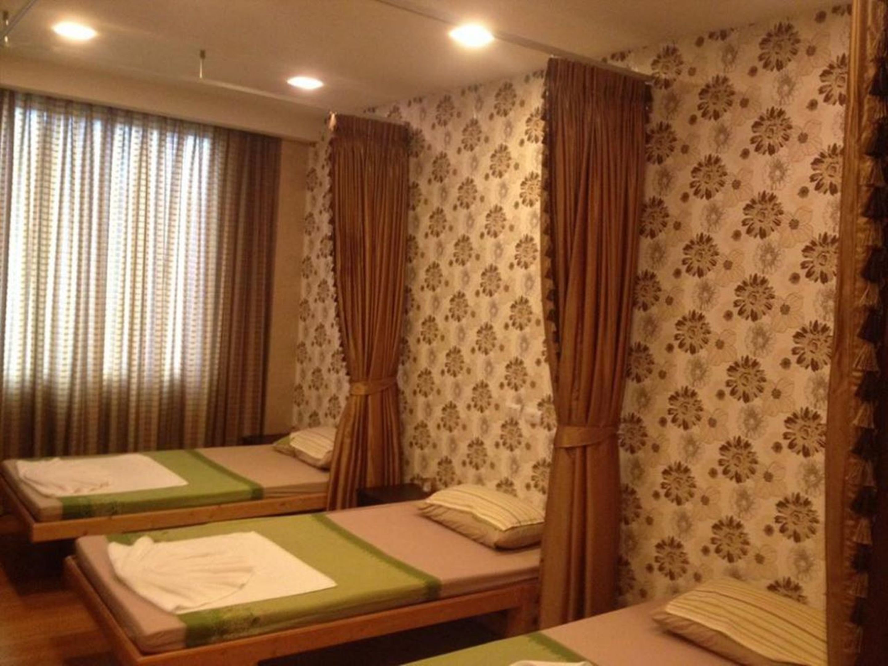 A room with three single beds, each with a green bedspread and white pillows. The walls have a floral pattern, and there are curtains on the windows.