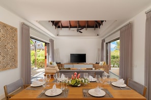 Un comedor con una mesa de madera preparada para una comida, un televisor al fondo y vistas al exterior.