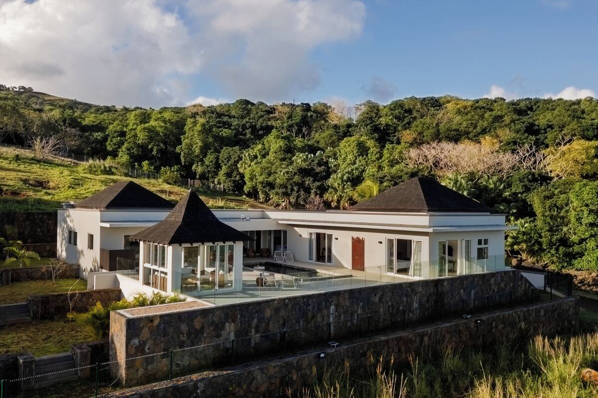 Une maison moderne avec une piscine, entourée d’une végétation luxuriante et de collines.