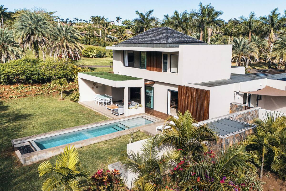 Une maison moderne à deux étages, dotée d’une piscine, entourée d’une végétation luxuriante et de palmiers.