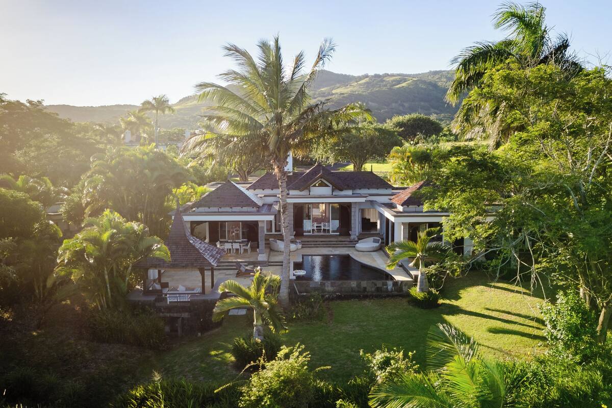 Une grande maison avec une piscine, entourée d’une végétation luxuriante et avec des collines en arrière-plan.