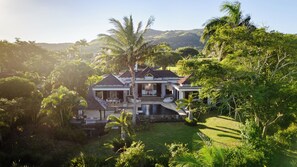 Une grande maison avec une piscine, entourée d’une végétation luxuriante et avec des collines en arrière-plan.