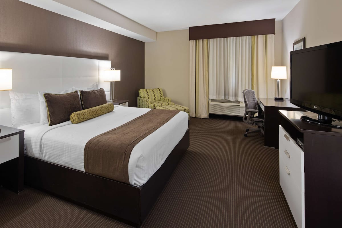 A hotel room with a large bed, two bedside tables with lamps, a desk with a TV, a chair, and an air conditioning unit.