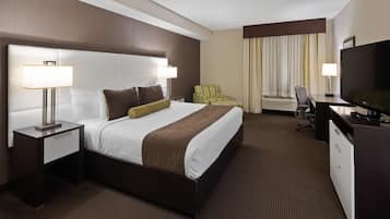 A hotel room with a large bed, two bedside tables with lamps, a desk with a TV, a chair, and an air conditioning unit.