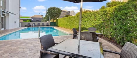 A poolside area with a rectangular glass table and wicker chairs, surrounded by a hedge and a building in the background.
