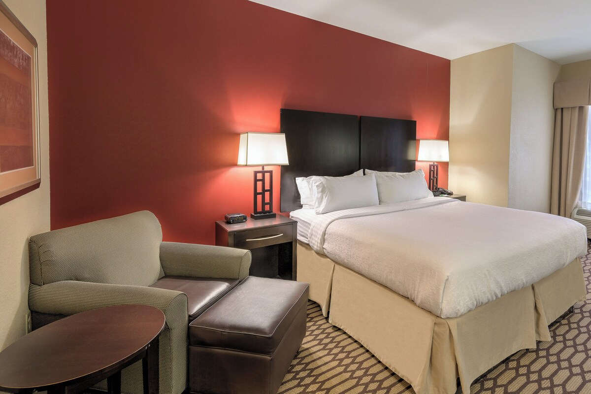 Habitación de hotel con cama, dos mesitas de noche, una silla, una pequeña mesa y una ventana con cortinas.