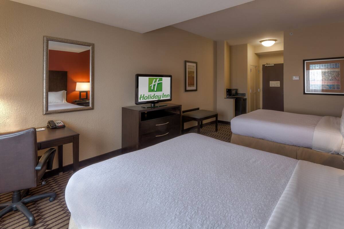 A hotel room with two beds, a desk with a computer, a telephone, and a television displaying the Holiday Inn logo.