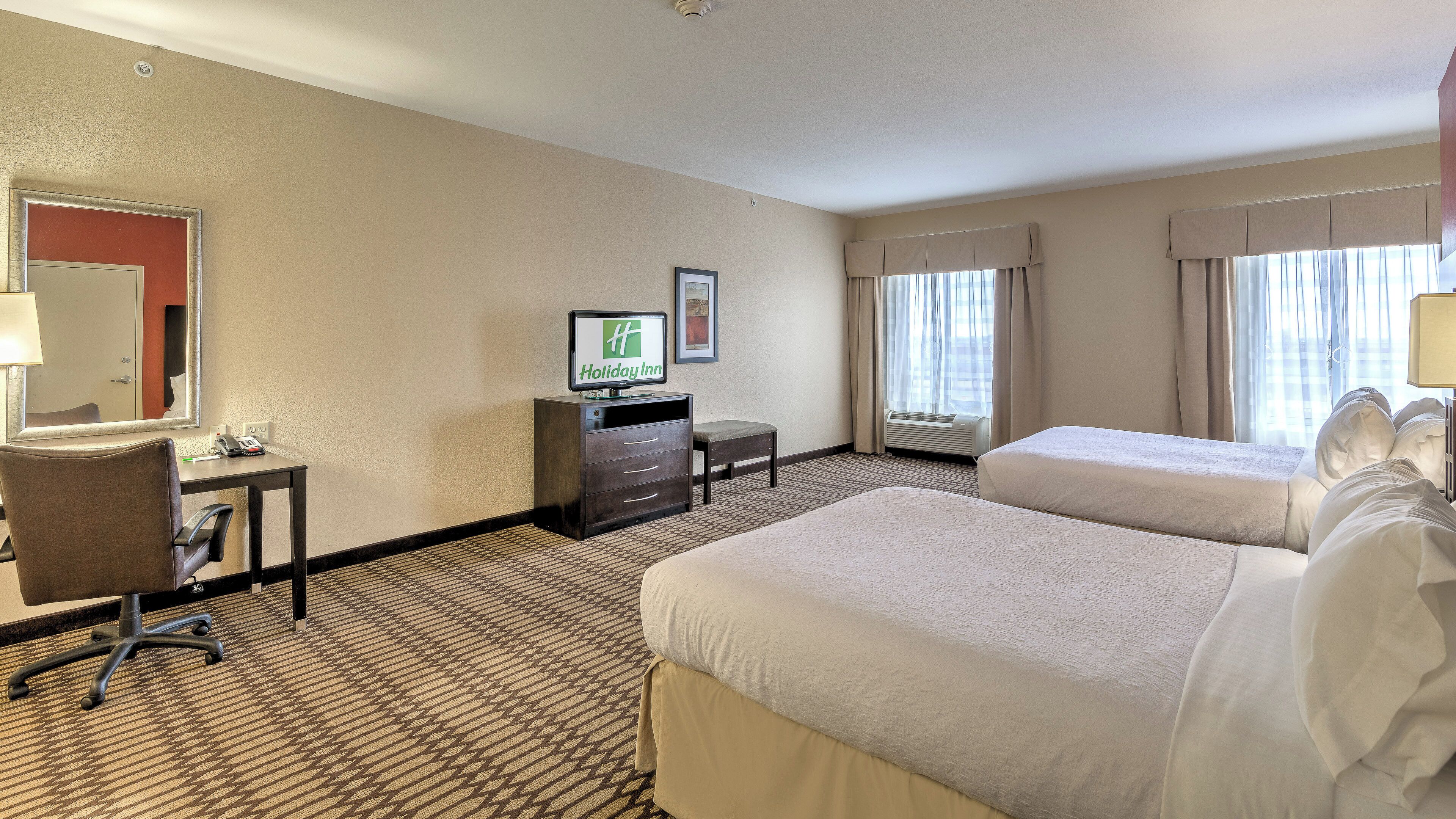 A hotel room with two beds, a desk with a computer, a chair, a TV, and a window with curtains.