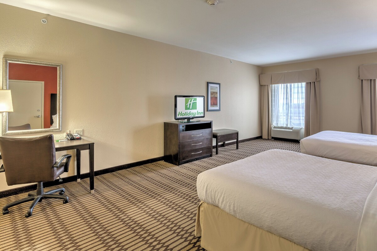 Habitación de hotel con dos camas, un escritorio con computadora, una silla, un televisor y una ventana con cortinas.