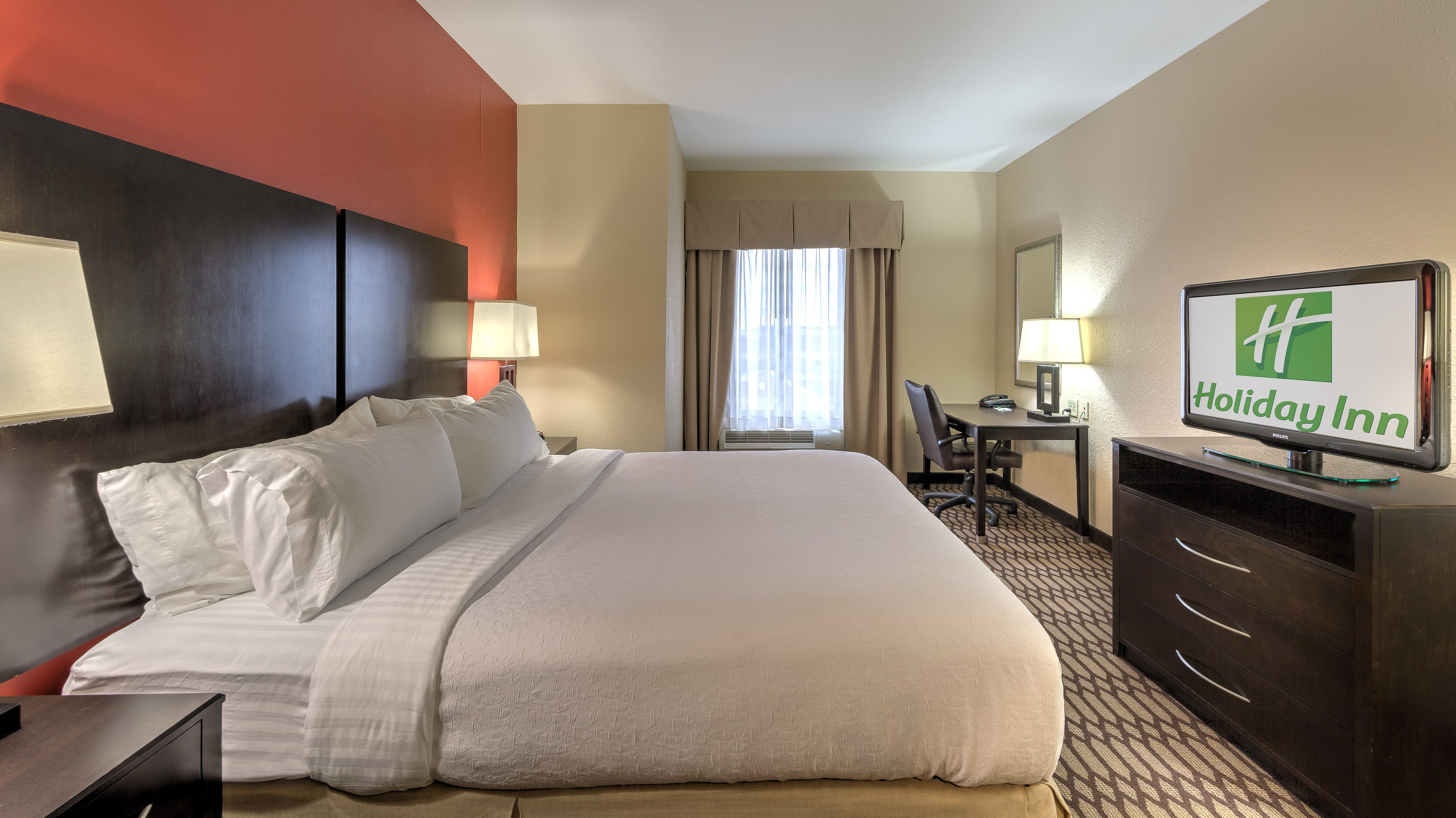 A hotel room with a large bed, a desk, a chair, a television, and a window with curtains.