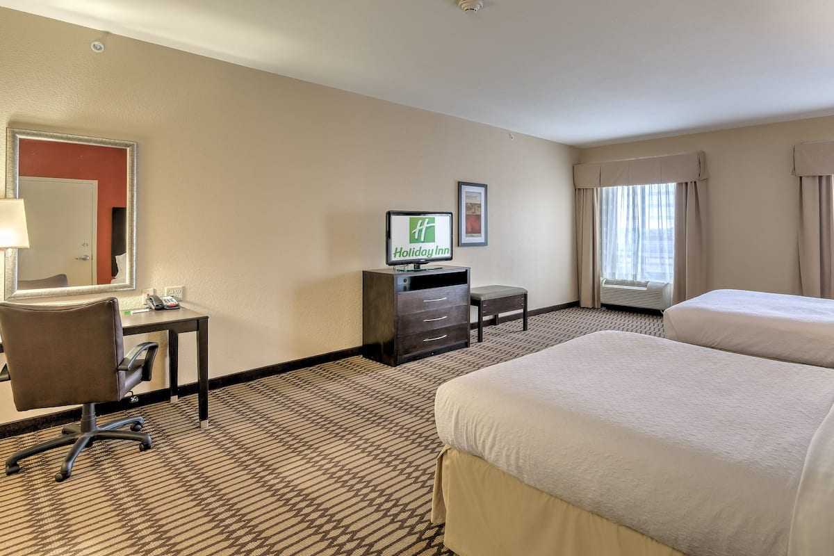A hotel room with two beds, a desk with a chair, a TV, and a large window with curtains.